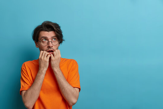 Scared nervous man bites finger nails and looks upwards with anxious expression, suffers phobia, terrified about problems, wears glasses and orange t shirt, isolated on blue background, empty space