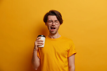 Tired male employee feels fatigue and exhausted, holds takeout coffee, yawns and wants to sleep, stressed of overtime job, wears glasses, stands with closed eyes, isolated on bright yellow background