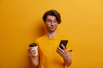 Handsome guy with funny expression, chats in cellphone, reads strange received message, drinks hot beverage from disposable cup, connected to wireless internet, dressed in casual wear, yellow color