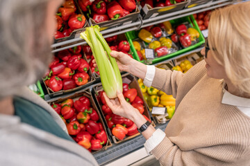 Mature female in beige knitted sweater holding fresh celery in supermarket