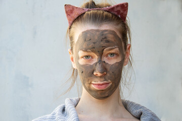 A light-skinned young woman put a clay mask on her face with freckles. It stands near a heterogeneously painted blue wall. On the head is wearing a red bezel with ears