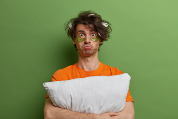 Fototapeta premium Displeased sullen man embraces pillow, looks with unhappy sleepy expression, wakes up early to have beauty treatments, wears under eye pads for removing wrinkles and fine lines, cares about complexion
