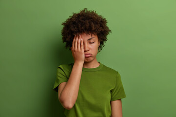 Fototapeta na wymiar Tired exhausted woman makes face palm, has problems with health, sleepy displeased expression, sighs from tiredness, wears casual green t shirt, poses indoor. Feeling fatigued and overworked
