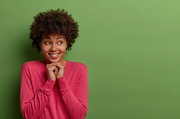 Fototapeta na wymiar Good looking pleased ethnic curly woman has good mood and expresses positive emotions, keeps hands under chin, looks aside, has toothy smile, dressed in rosy jumper, isolated on green background
