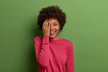Horizontal shot of positive dark skinned woman makes face palm, chuckles and covers half of face, expresses joy, wears rosy jumper, poses against green background. Positive emotions concept.
