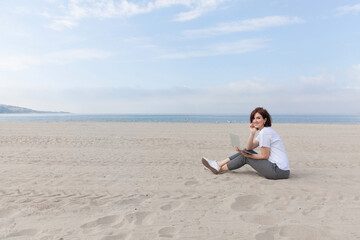 Fototapeta na wymiar Smiling pretty woman sitting on the beach with her laptop. Freelance concept.