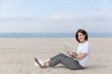 Smiling pretty woman sitting on the beach with her laptop. Freelance concept.