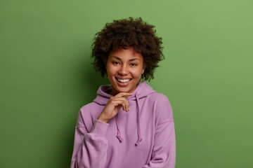 Indoor shot of happy dark skinned woman with Afro hair, keeps hand under chin, shows white teeth,...