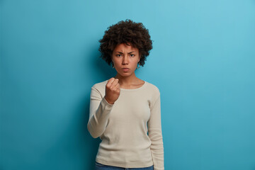 Fototapeta na wymiar Outraged furious woman with Afro hair, clenches fist with anger, experiences strong aggression, smirks face, wears casual white long sleeve jumper, expresses irritation, stands against blue wall