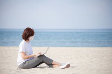Young woman in medical mask sitting with laptop on empty coast, protective equipment during coronavirus. Freelance concept, working on the beach. Distance education on the beach with medical mask.