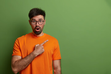 Look, advertise here! Speechless impressed man points at upper right corner, demonstrates empty space for promo or commercial presentation, wears orange t shirt and spectacles, isolated on green wall