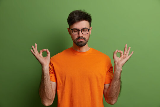 Mindful Calm Bearded Young Man Practices Yoga, Breathes Deeply, Enjoys Peaceful Meditation, Keeps Eyes Closed, Wears Orange T Shirt, Isolated On Green Background. Zen Concept. Time For Yoga.