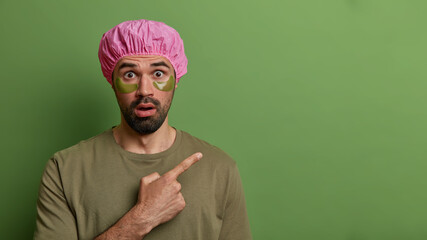 Horizontal view of shocked bearded man points index finger on empty space, has bugged eyes, applies collagen patches for reducing wrinkles or puffiness, isolated on green background takes care of skin