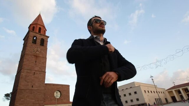 Ragazzo in giacca e occhiali da sole si diverte a ballare in in una piazza di una citt&agrave; con un campanile sullo sfondo in una bella giornata con il cielo azzurro