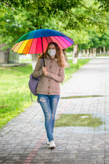 Caucasian girl in a protective mask walks under an umbrella on an empty street in spring rain. Safety and social distance during a coronavirus pandemic. New normal, the implications of quarantine