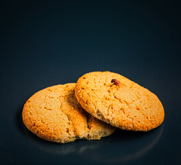 Two oatmeal cookies lie on top of each other and one of them is bitten. The photo is on a black background with reflections.
