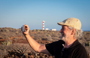 Smiling retired senior man with cap and a black polo shirt while taking a selfie. A lighthouse and the blue sea in the background, arid tropical area with volcanic rocks.