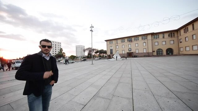 ragazzo moro con giacca e occhiali scuri, gira per una piazza nel centro di una citt&agrave;
