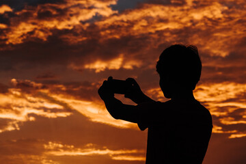 Silhouette of a kid using mobile phone in front of the sky during sunset