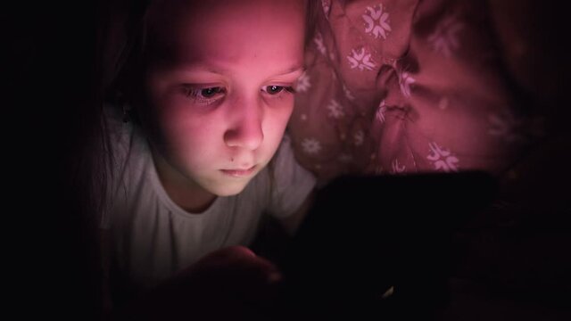 A young little girl is hiding under a blanket to use a digital tablet smartphone device late after sleep. Loneliness of young children and rescue on the phone.
