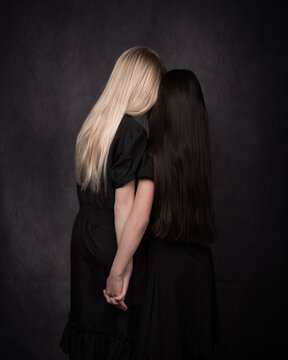 Blonde and black haired girls in studio portrait classic renaissance style seen from back holding hands