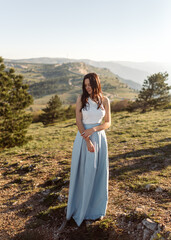 Fototapeta na wymiar Beautiful bride in white blouse and blue skirt standing on the picturesque hill in the sunlight. An attractive girl in a minimalistic dress with jewelry rings on her hands stands in a green field.