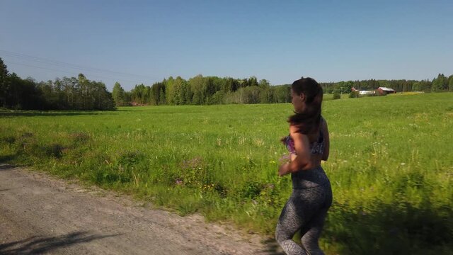 A young fit woman in leggings and a sports bra top is jogging in the Swedish countryside during a sunny summer day by herself. Cute red wooden houses in the surroundings.
