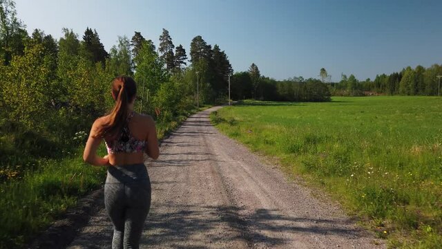 A young fit woman in leggings and a sports bra top is jogging in the Swedish countryside during a sunny summer day by herself on a gravel road. Green and lush fields and forest in the background.