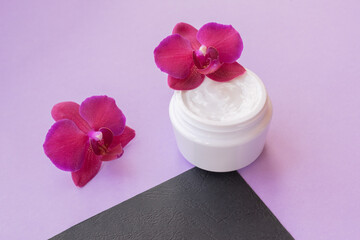 White container with cream for face and body with two magenta colored orchid flowers on purple and black background. Concept of delicate or eco friendly cosmetics