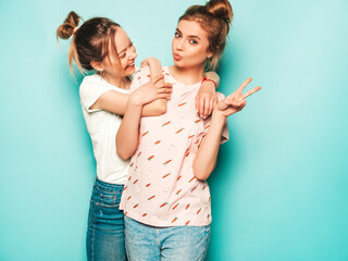 Two young beautiful blond smiling hipster girls in trendy summer hipster jeans clothes. Sexy carefree women posing near blue wall. Trendy and positive models having fun