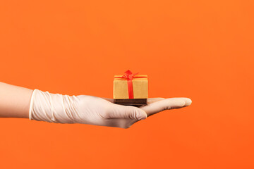 Profile side view closeup of human hand in white surgical gloves holding small gift box. sharing, giving wedding proposal, or delivery concept. indoor, studio shot, isolated on orange background.