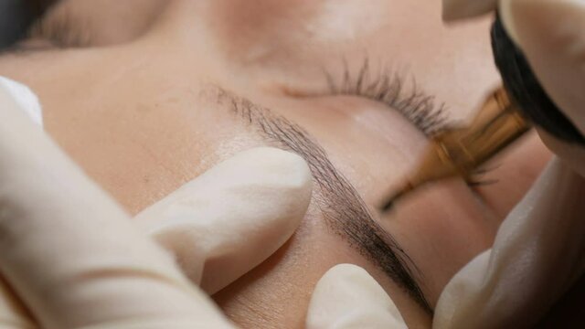 Beautician in black gloves makes permanent makeup correction of the shape of the eyebrows to a young beautiful girl. Microblading, eyebrow tattoo in a beauty salon.