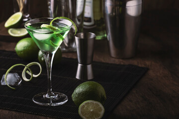 Green martini alcoholic cocktail in glass with dry gin, vermouth, liquor, lime zest and ice, steel bar tools, dark background