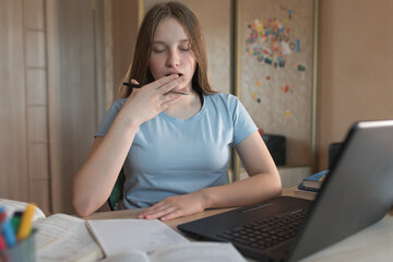 Teenager girl, tired yawns, malaise, sleep, break and lethargy at school. E-education and online lessons, distance learning. Laptop and abstracts on the training table at home.