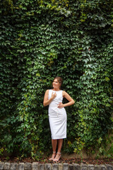 Young woman weared in white simple wedding dress on background of greenery wall. Fashion style girl happy bride weared in pencil-dress. Street style Woman portrait. Modern urban wedding walk
