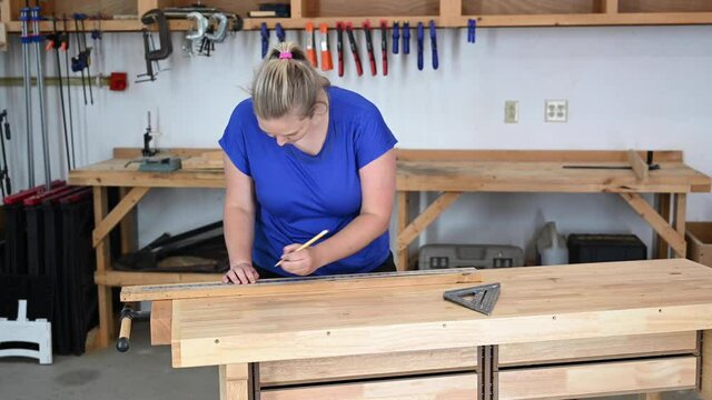 Female caucasian young adult woodworker measuring and marking wood with a metal yardstick and triangle.