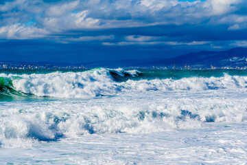 Storm waves in white foam rush in rows along the Tsemesskaya Bay. Blue sky and green sea. Dangerous and dramatic. In the background, mountains, multi-storey buildings of the city and port.