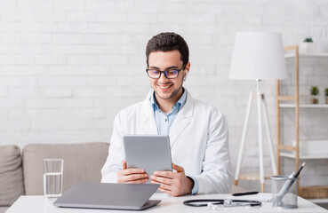 Online visit to doctor. Man looks at tablet, sitting at table with laptop