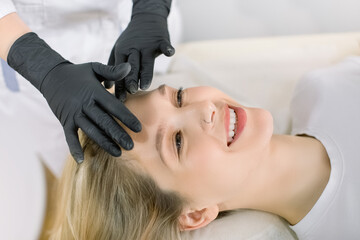 Obraz na płótnie Canvas Cropped image of hands of female professional cosmetologist making face massage to young smiling woman feeling relaxed in beauty salon. Concept of antiaging facial skin treatments and massage