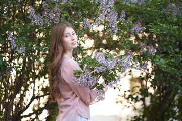A young girl with light long wavy hair, standing in the leaves of trees on the girl's cheeks lilac petals, she looks directly into the camera   