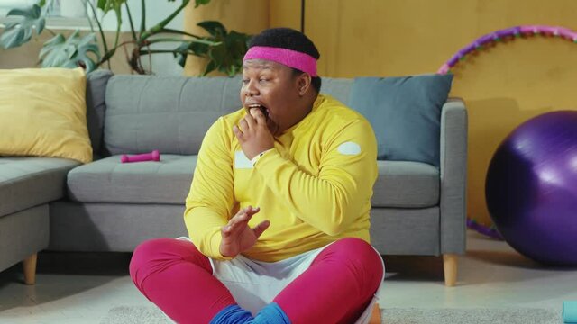 Silly overweight black man sitting in lotus pose and eating cookie without permission during meditation session with personal coach. Concept of fun, obesity, junk food.