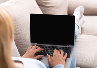 Work online. Woman typing on laptop with blank screen, lying on sofa in home interior, shoulder view