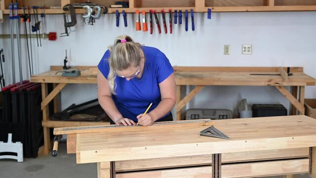 Female caucasian young adult woodworker measuring and marking wood with a metal yardstick and triangle.
