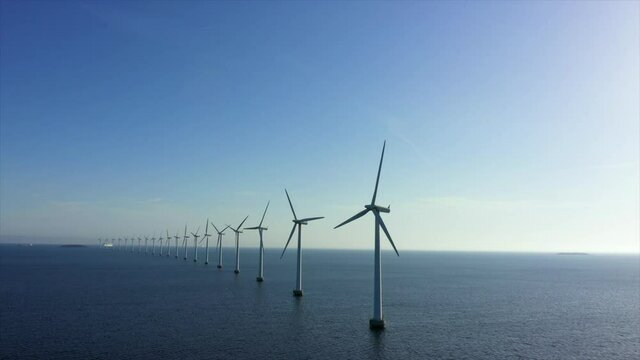 Aerial panning shot of wind turbines in sea against blue sky, drone flying over water on sunny day - Copenhagen, Denmark
