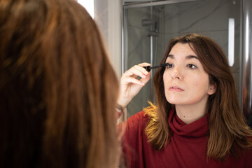 Attractive young female putting eye-mascara in her right eye in front of a mirror on a modern bathroom