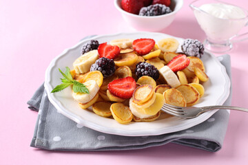 Trendy tiny pancakes for breakfast with strawberry, banana and frozen blackberry in white plate on pink background. Closeup