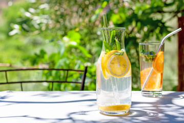 Cold refreshing summer drink or cocktail in carafe with glass straw. Detox water or lemonade with slices of lemon and basil in glass carafe with recyclable straw. Summertime zero wast concept.