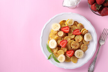 Trendy tiny pancakes for breakfast with strawberry, banana and sour cream in white plate on pink background. View from above. Copy space for text or design
