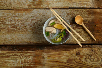A light plate of Vietnamese Pho soup with chicken on a wooden table next to lemon onion and garlic.