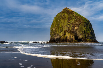 Naklejka premium Haystack Rock reflected in the wet sand, with wave foam and blue sky and clouds, Canon Beach, Oregon